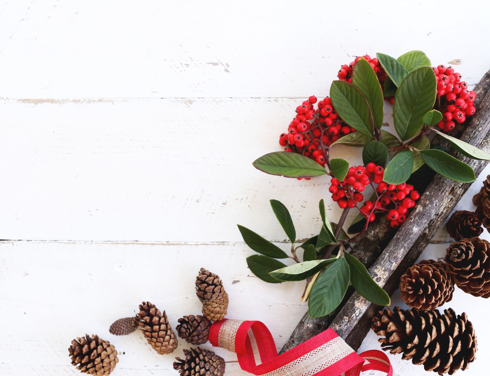 Top-down view of red berries, pine cones, and festive ribbon on a rustic wooden surface.
